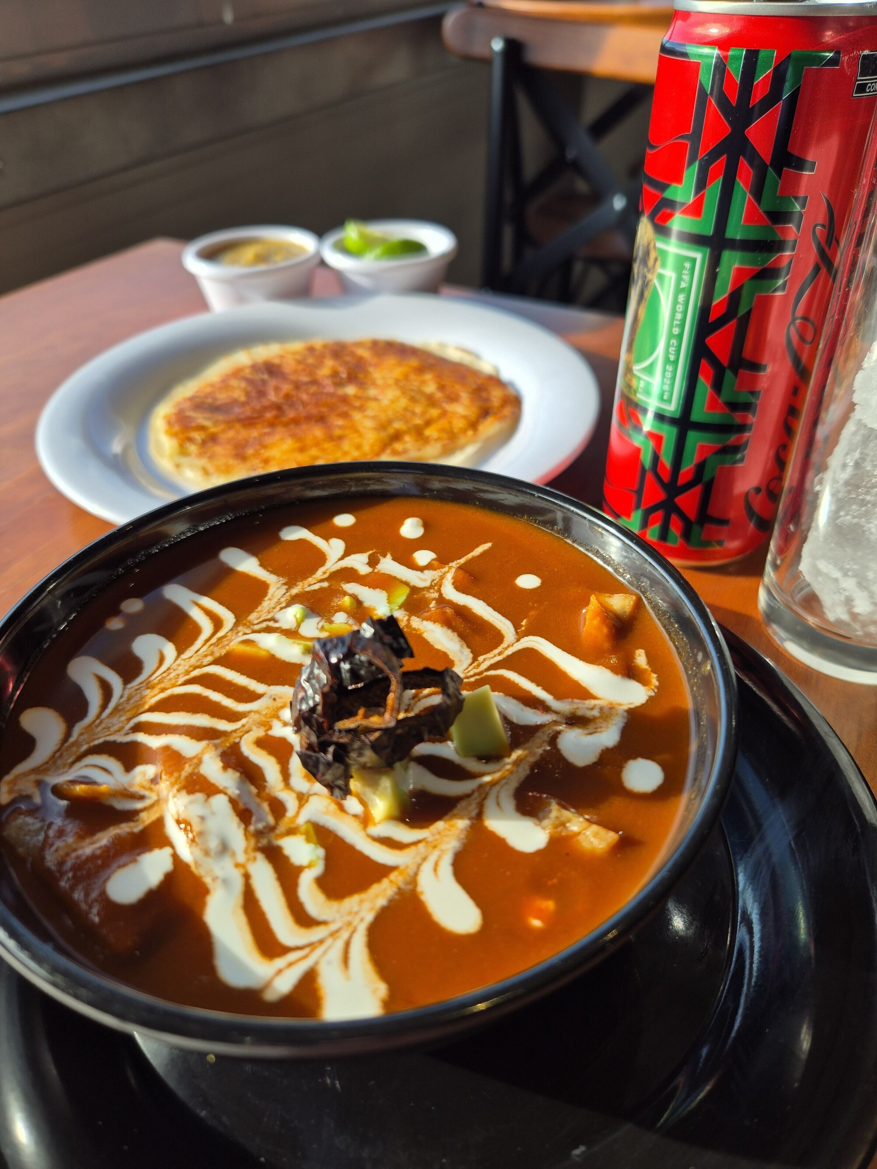 A bowl of soup with a garnish, a plate with a food item, a can of drink, and a glass of water on a table.