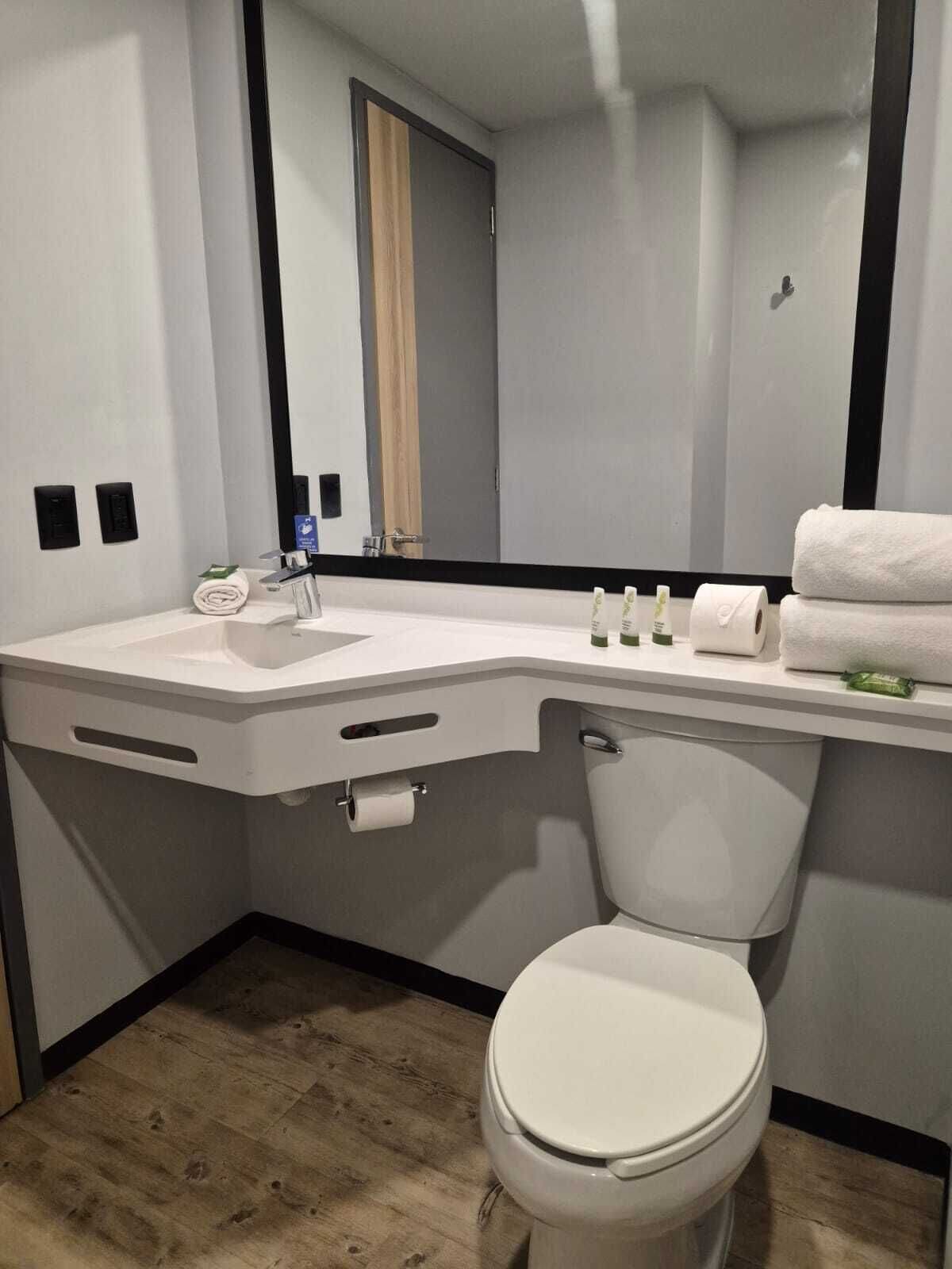 Een moderne badkamer met een witte wastafel, toilet en spiegel. Er staan toiletartikelen op de aanrecht en handdoeken gestapeld aan de muur.