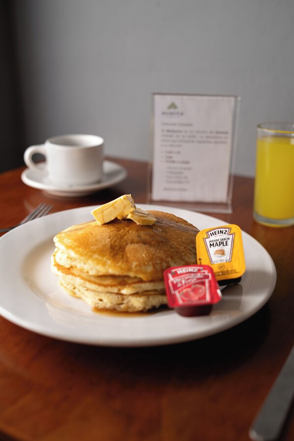 Una pila de tortitas con mantequilla y jarabe de arce, una taza de café y un vaso de jugo de naranja sobre una mesa de madera.
