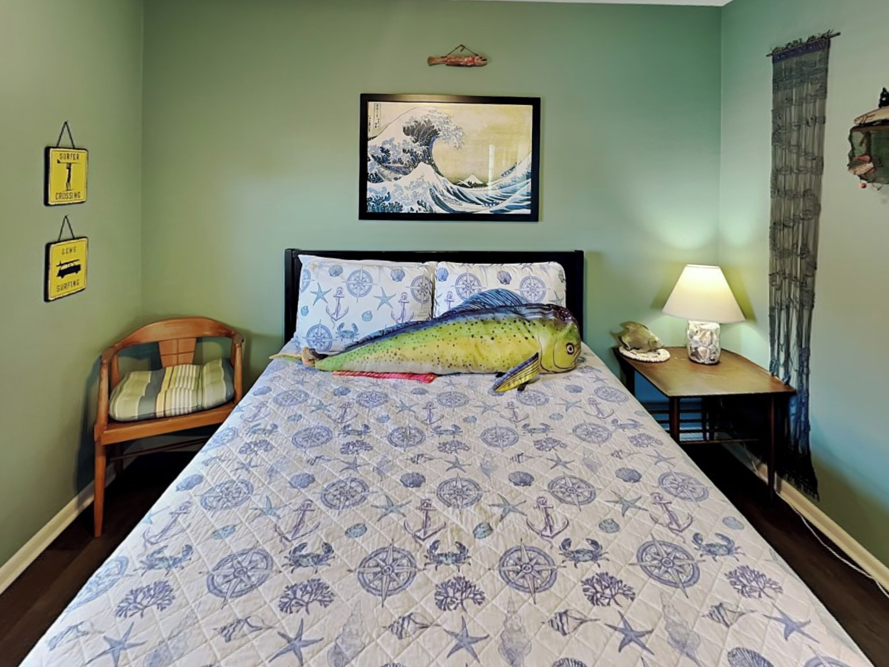 A bed with a nautical-themed bedspread, a framed artwork above the bed, a chair to the left, and a nightstand with a lamp to the right.