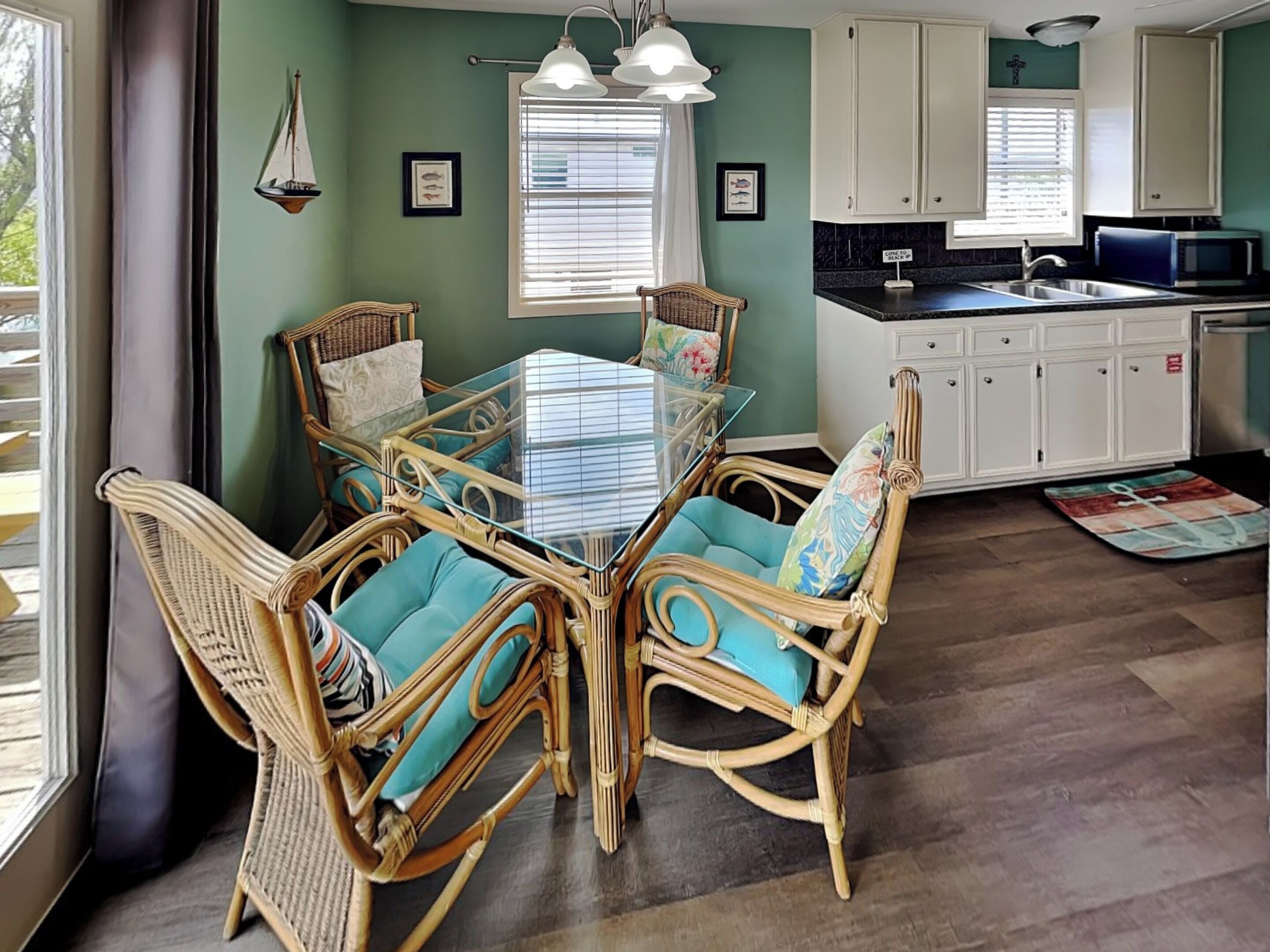 Un coin repas avec une table à plateau de verre entourée de chaises en osier dotées de coussins turquoise, une cuisine avec des meubles blancs et une fenêtre équipée de stores.