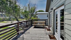 A wooden deck with a railing, picnic table, and chairs, overlooking a road and trees. - The Anchorage by Silver Sands Vacation Rentals (Port Aransas)