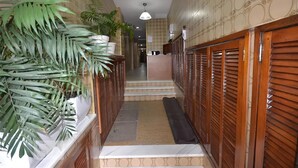Un estrecho pasillo con una escalera que sube a un piso superior, flanqueado por contraventanas de madera y plantas en macetas.