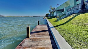 Eine hölzerne Pier erstreckt sich über ruhiges Wasser, neben einem Gebäude mit grüner Fassade und einer Rasenfläche.