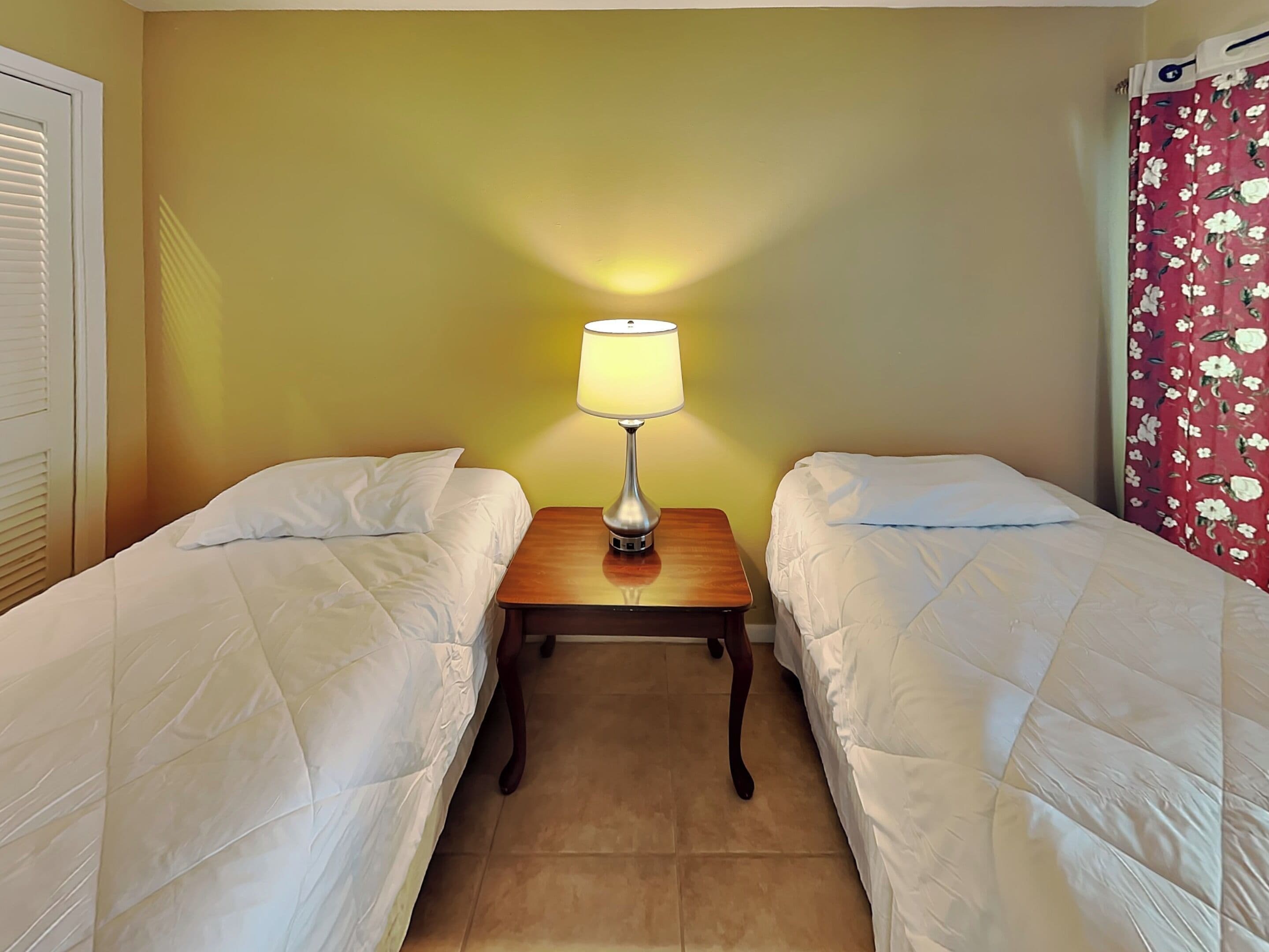 Habitación de hotel con dos camas individuales, una mesita de noche con lámpara y una cortina floral roja.