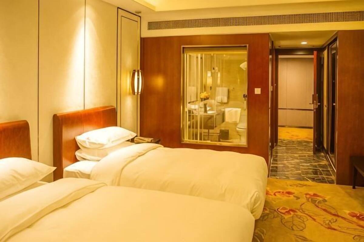 A hotel room with two beds, a bathroom with a glass door, and a hallway leading to other rooms.