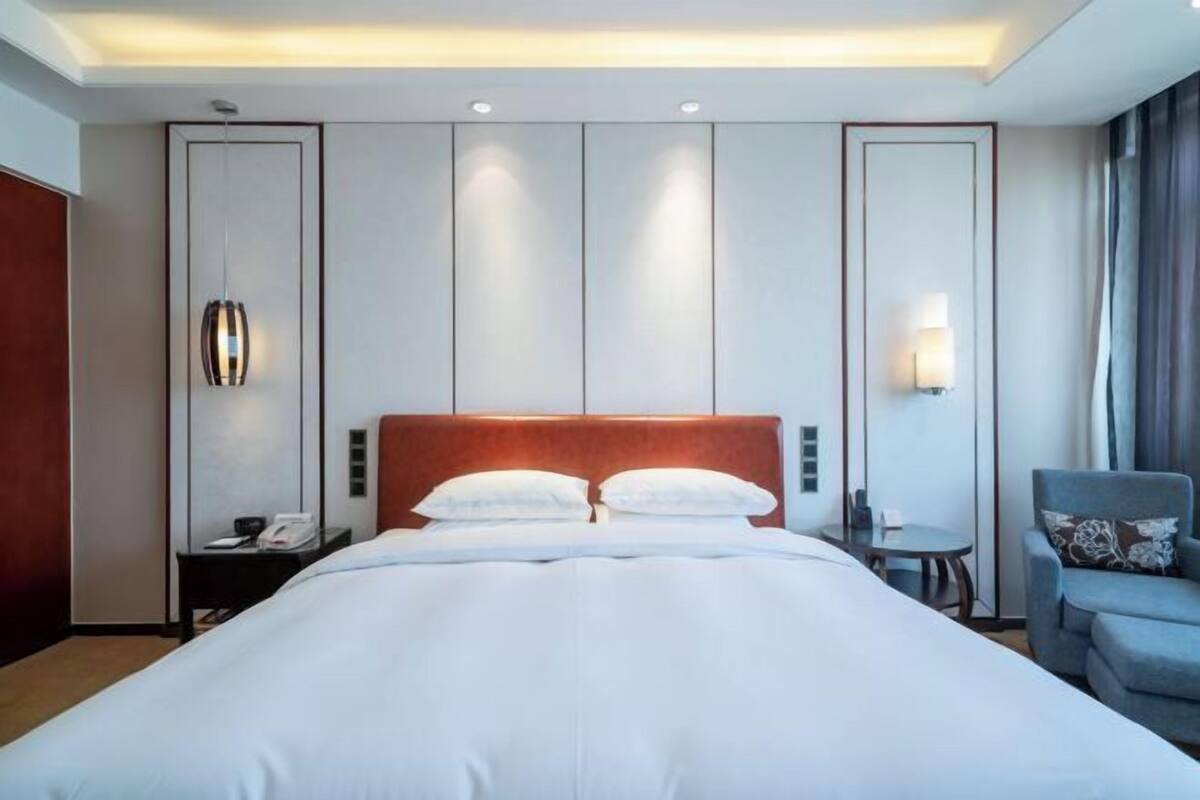 A hotel room with a large bed, a bedside table with a lamp, a small table with a chair, and a view of the outside.