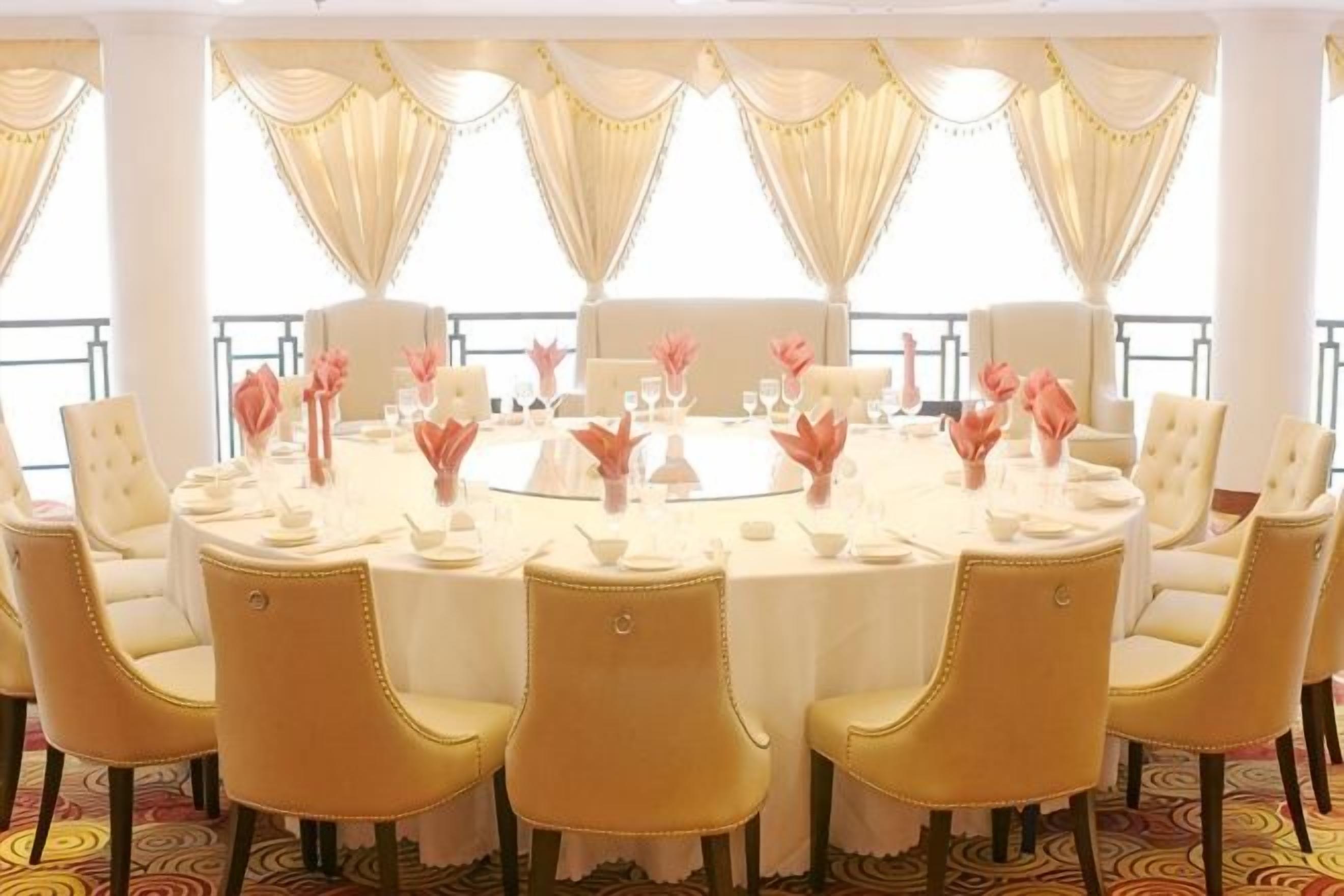 A round dining table with chairs, set for a meal, in a room with large windows and curtains.