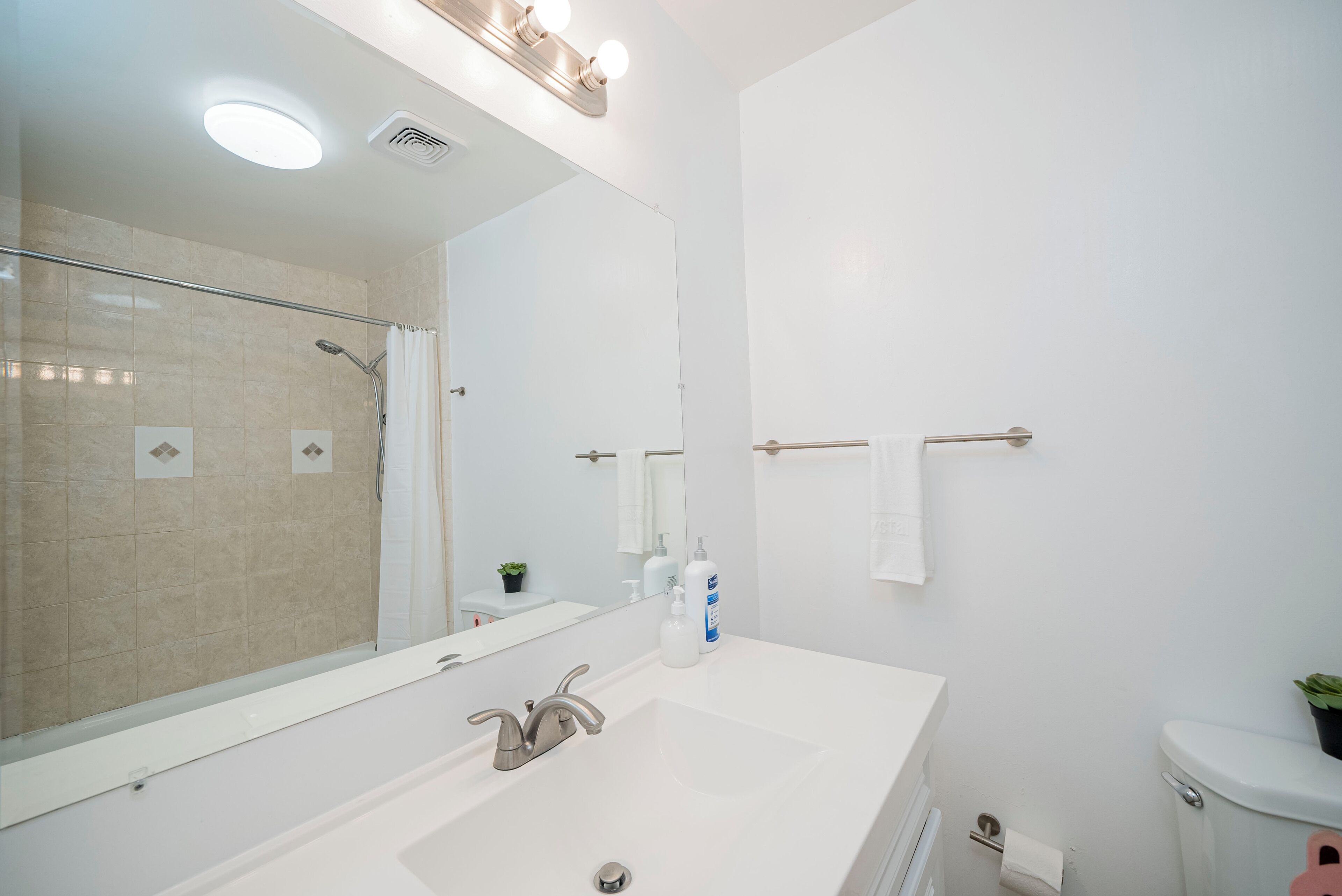 A modern bathroom with a large mirror, a shower, a sink with two bottles of soap, and a toilet.