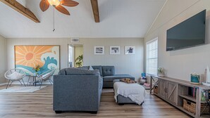 Un salon avec un canapé gris, un petit pouf, un ventilateur de plafond et un téléviseur fixé au mur.