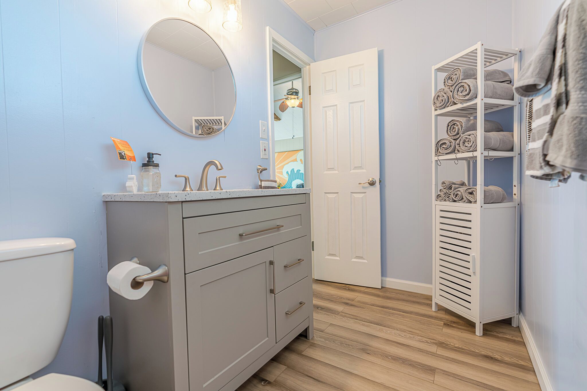 A bathroom with a grey vanity, a round mirror, a towel rack, and a storage unit with towels.