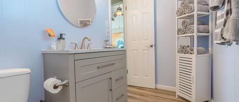 A bathroom with a grey vanity, a round mirror, a towel rack, and a storage unit with towels.
