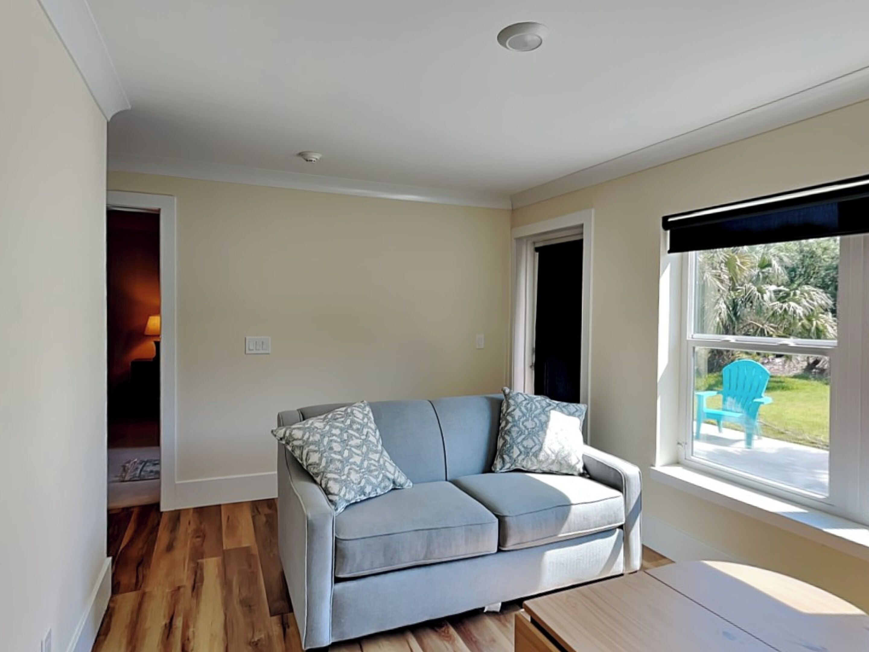 A living room with a grey sofa, patterned cushions, a wooden coffee table, and a view of a garden through a window.