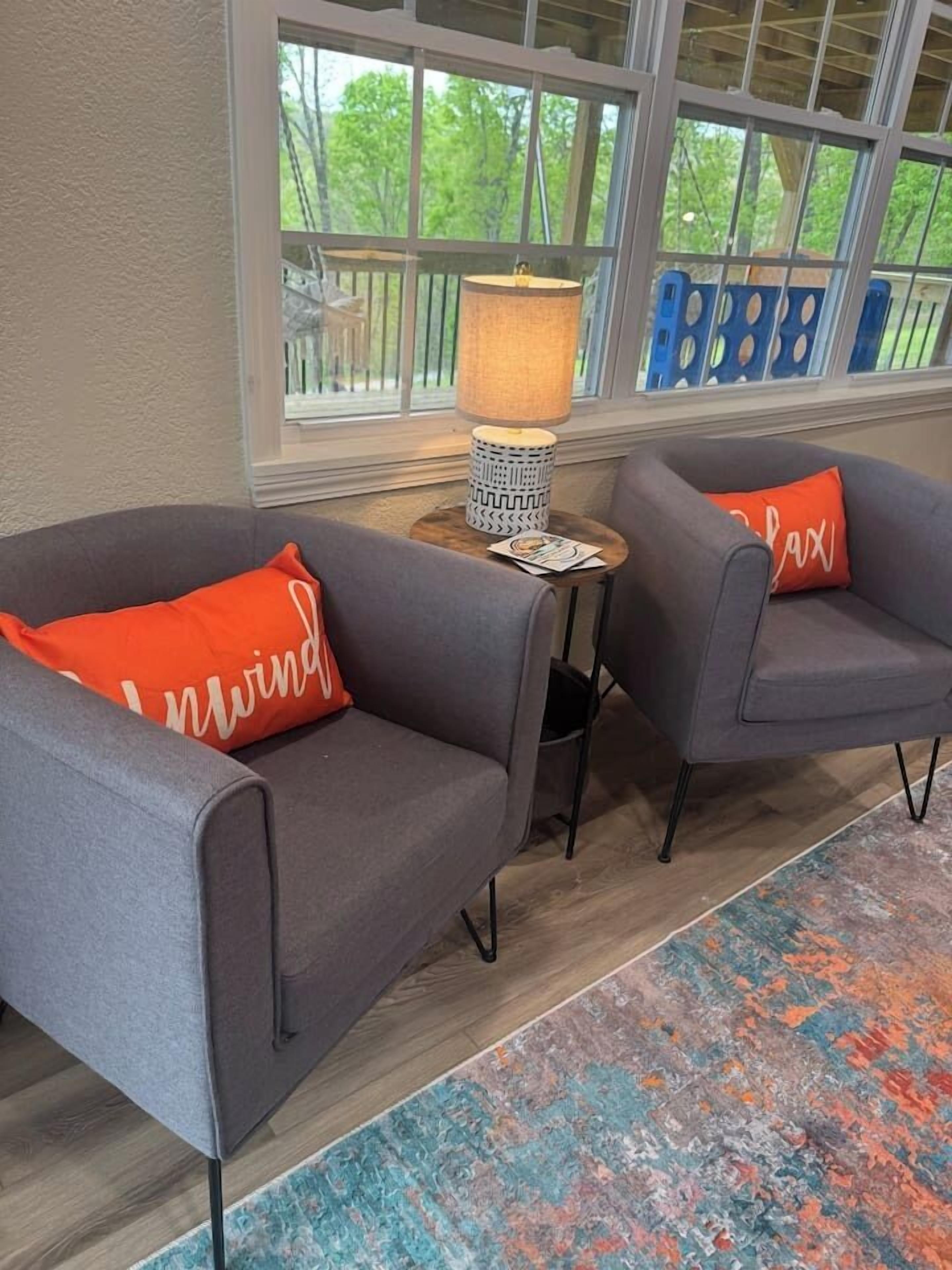 A room with two grey armchairs, a small round table with a lamp, and a colorful rug.