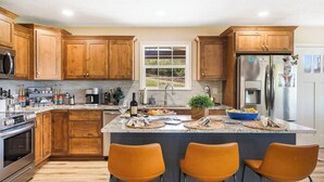 A modern kitchen with wooden cabinets, stainless steel appliances, a large island with bar stools, and a view of the outdoors through a window. - The Breakaway at Table Rock- Sauna, Hot Tub, Kayak, Full Upper and Lower Porches (Omaha)