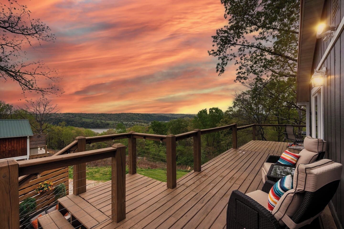 Una terraza de madera con barandilla, una silla con cojines y vistas a un paisaje con árboles y un atardecer.