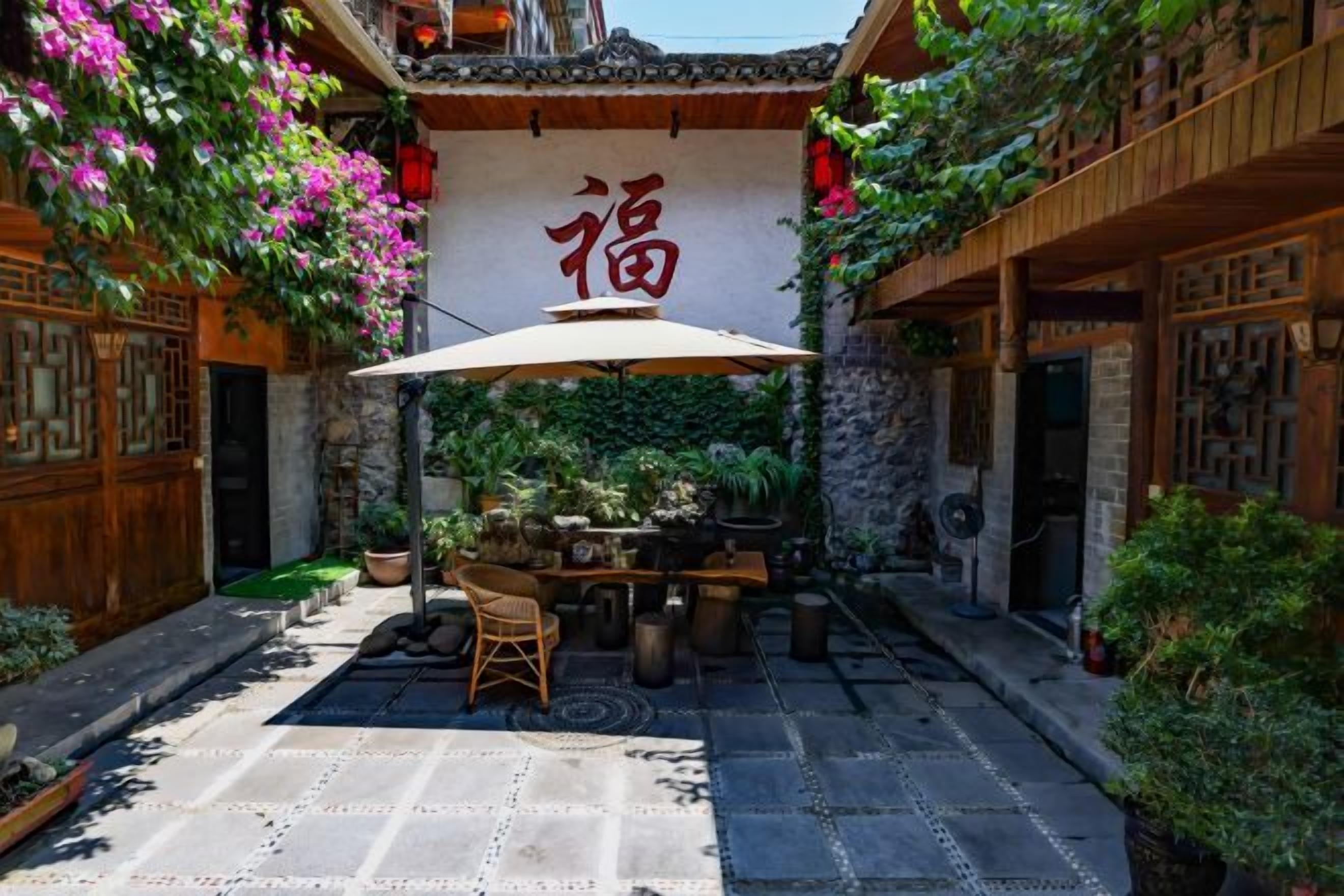 A courtyard with a table and chairs under a large umbrella, surrounded by potted plants and a traditional Chinese wall with a sign.