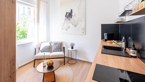 A modern living room with a sofa, a round table with a bowl of fruit, a painting on the wall, and a kitchen area with a coffee machine and a bottle. - Apartment in Koblenz Near Deutsches Eck (Koblenz)