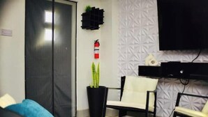 A modern living room with a sofa, a coffee table, a white chair, a potted plant, a fire extinguisher, and a mounted television. - Ideal Studio Apartment,  AC, WIFI & parking, minutes from Stadium and the city (Mount Gay)
