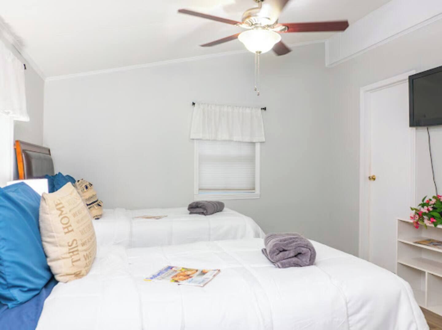 A bedroom with two beds, a ceiling fan, a window with a white valance, a TV on the wall, and a small shelf with flowers.