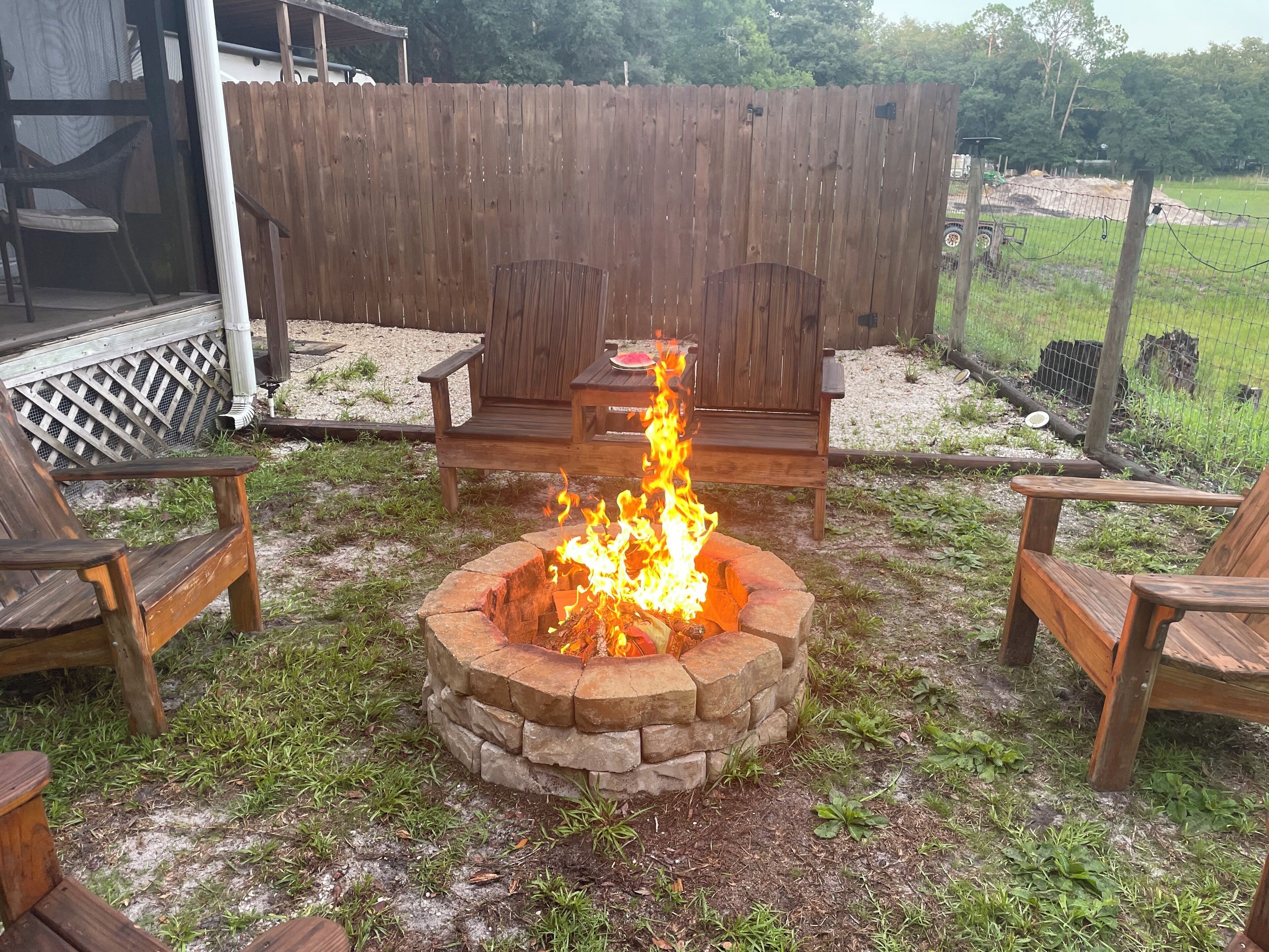 A fire pit surrounded by wooden chairs and a bench, with a wooden fence and greenery in the background.