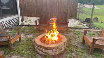 A fire pit surrounded by wooden chairs and a bench, with a wooden fence and greenery in the background.