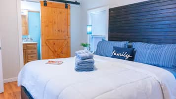 A bedroom with a large bed, a wooden door, a window with blinds, and a kitchen area visible through an open door.