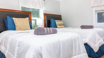 Two twin beds with white bedding and blue accents, a window with white curtains, and a wooden headboard.
