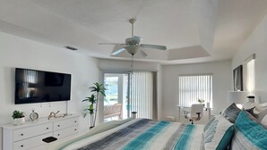 Een slaapkamer met een groot bed, een plafondventilator, een televisie, een bureau en een raam met jaloezieën.
