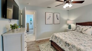 Une chambre à coucher comprenant un lit, un ventilateur de plafond, une télévision, une commode et une porte donnant sur une autre pièce.