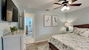 Une chambre à coucher comprenant un lit, un ventilateur de plafond, une télévision, une commode et une porte donnant sur une autre pièce.