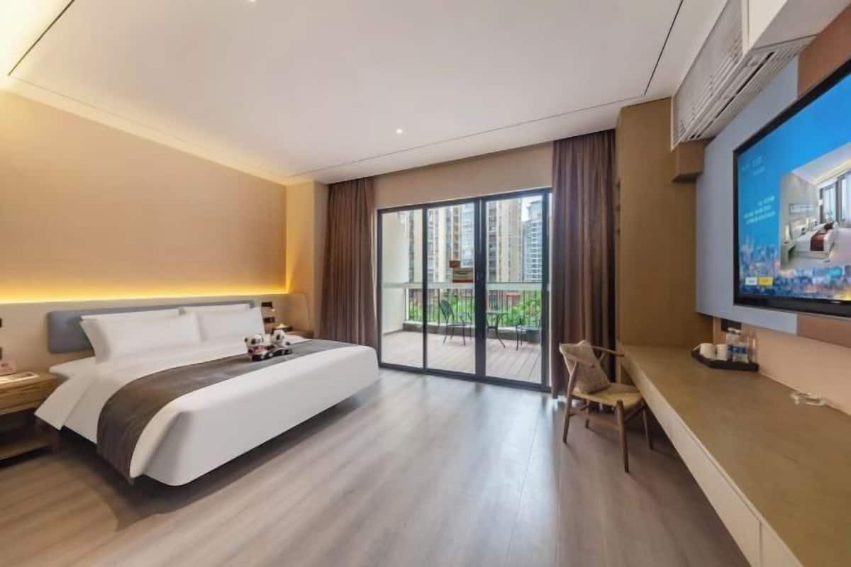 A modern hotel room with a large bed, a desk, a TV, and a balcony with a view of the city.