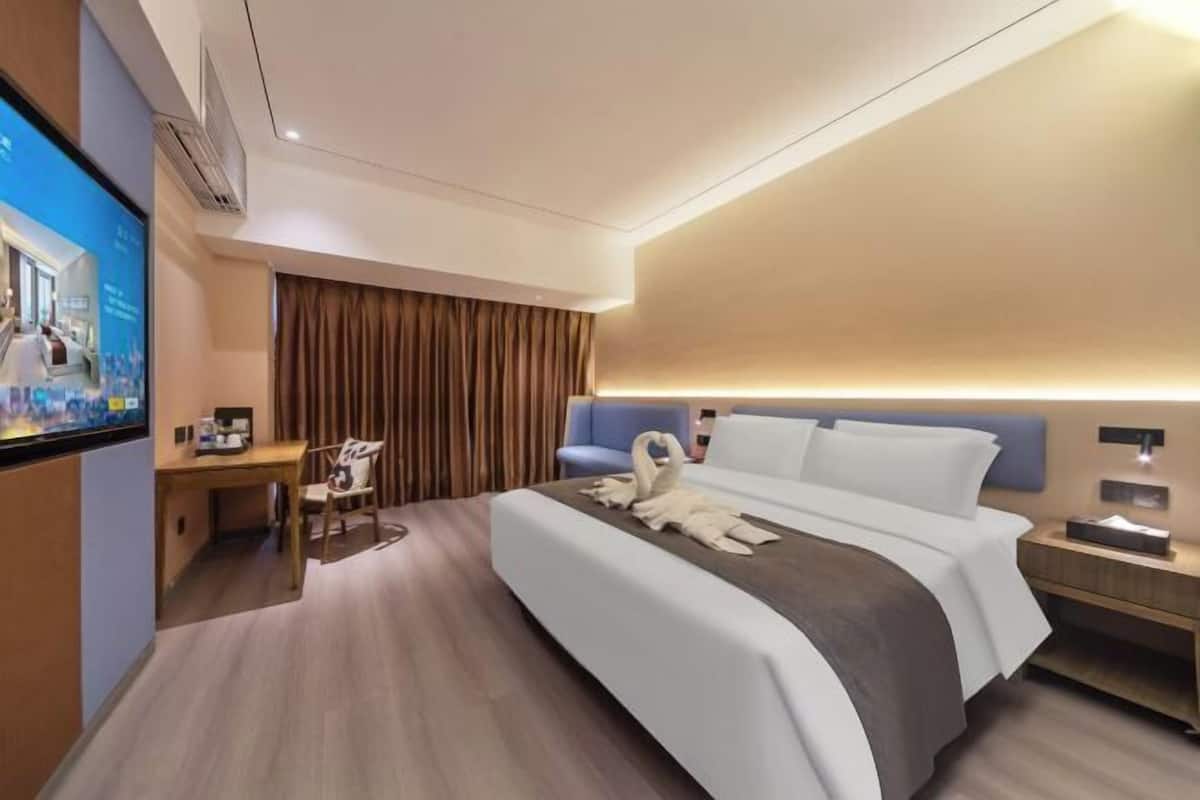 A modern hotel room with a bed, a desk, a chair, a TV, and a wall-mounted lamp.