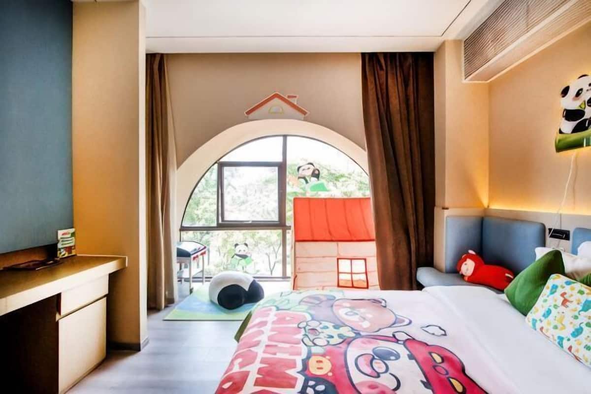 A hotel room with a bed, a desk, a window with curtains, and a view of greenery outside.