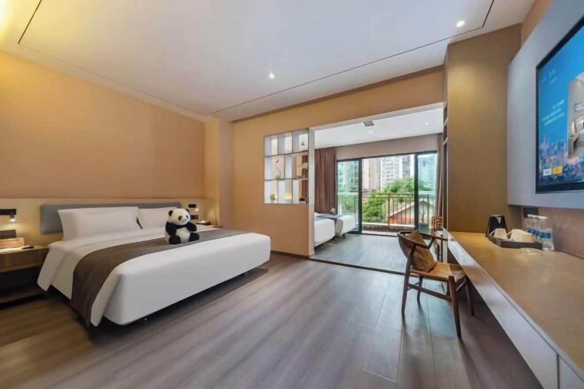 A hotel room with a bed, bedside table, a chair, a desk, and a large window with a view of buildings.
