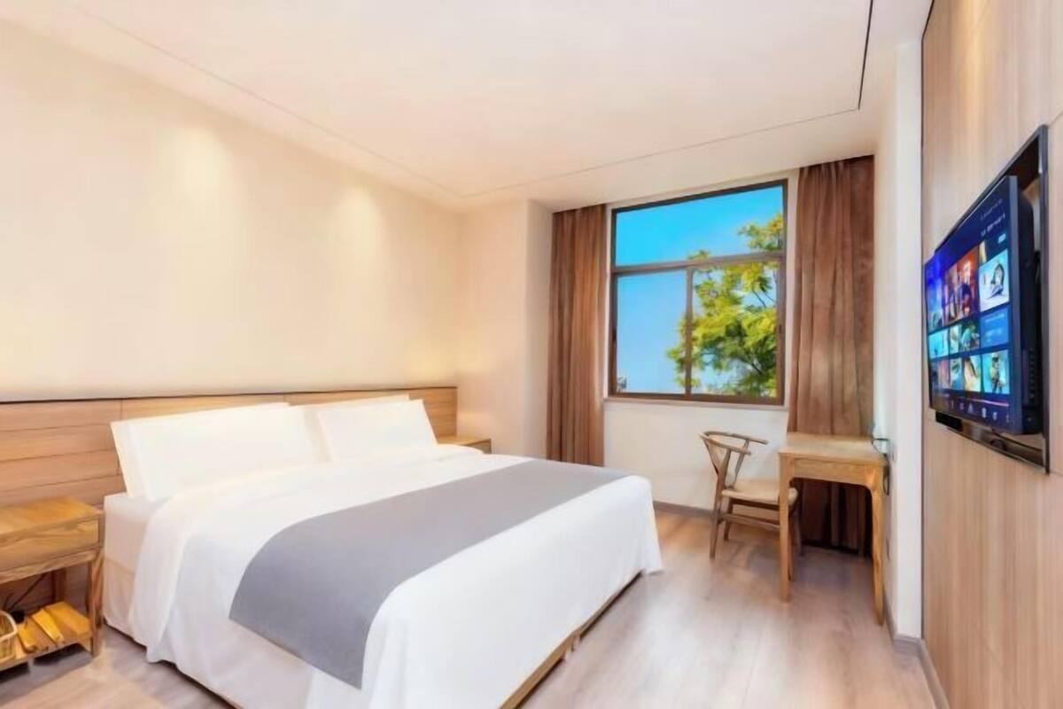 A hotel room with a bed, a desk, a chair, a TV, and a window with a view of trees.