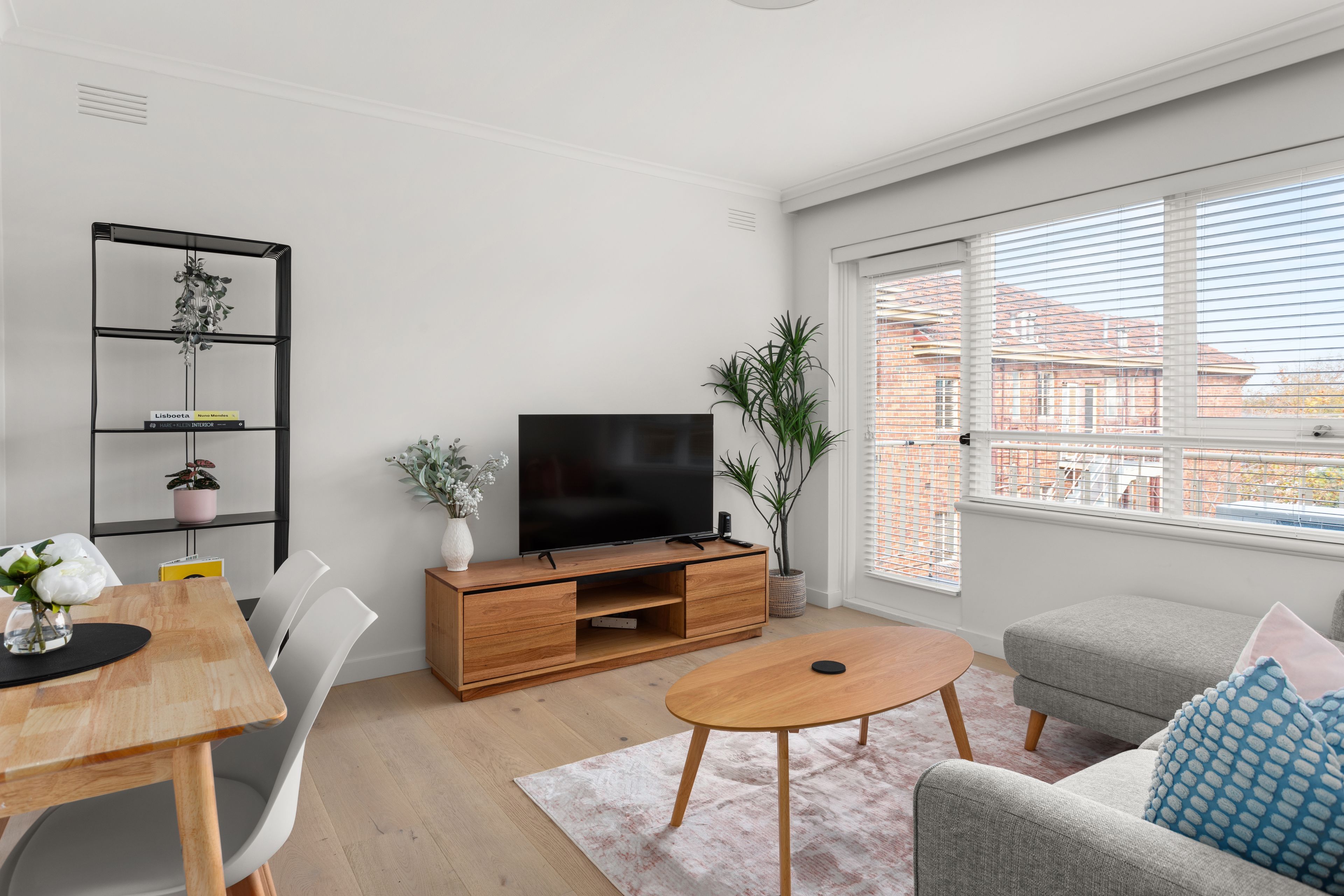 A modern living room with a flat-screen TV, wooden furniture, a dining table, and a large window with a view of buildings.