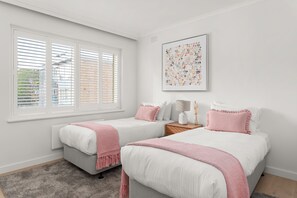 A bedroom with two beds, a window with blinds, a bedside table with a lamp, and a wall-mounted picture.