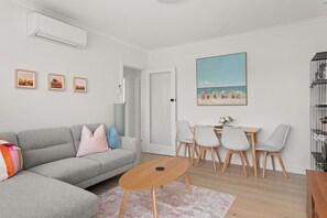 A modern living room with a grey sofa, a small round table, and a dining area with a white table and chairs. There are three framed artworks on the wall, a painting of a beach scene, and a door leading to another room.