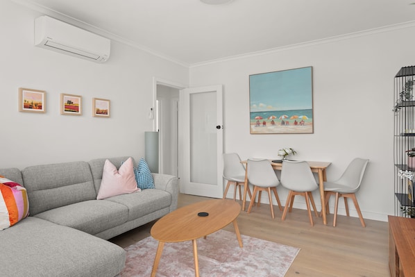 A modern living room with a grey sofa, a small round table, and a dining area with a white table and chairs. There are three framed artworks on the wall, a painting of a beach scene, and a door leading to another room.