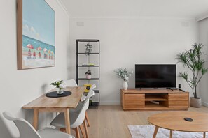 A modern living room with a wooden dining table, white chairs, a black TV stand, and a bookshelf.