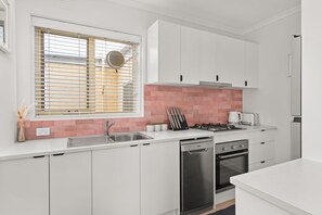 A modern kitchen with white cabinets, a stainless steel sink, a dishwasher, a stove, and a window with blinds.