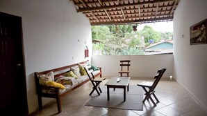 A room with a wooden ceiling, a window with a view of greenery, a sofa with cushions, and a small table with chairs. - POUSADA DOS GIRASSOIS TRANCOSO (Porto Seguro)