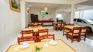 A restaurant with multiple tables and chairs, a television mounted on the wall, and a parking area visible through the open door. - POUSADA DOS GIRASSOIS TRANCOSO (Porto Seguro)