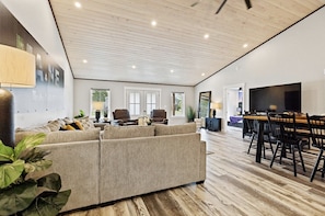 A spacious living room with a large sectional sofa, a dining area with a wooden table and chairs, and a flat-screen TV mounted on the wall.