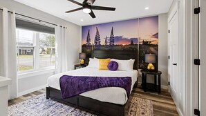 Una camera da letto con un letto, una ventola a soffitto, un comodino con una lampada, una finestra con tende e un affresco sulla parete.