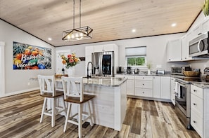 A modern kitchen with white cabinets, a central island with bar stools, a hanging light fixture, and a 'Welcome' sign.