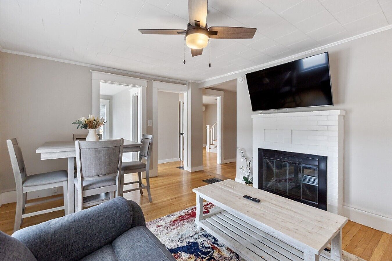 A living room with a fireplace, a television mounted above it, a dining area with chairs and a table, and a ceiling fan.