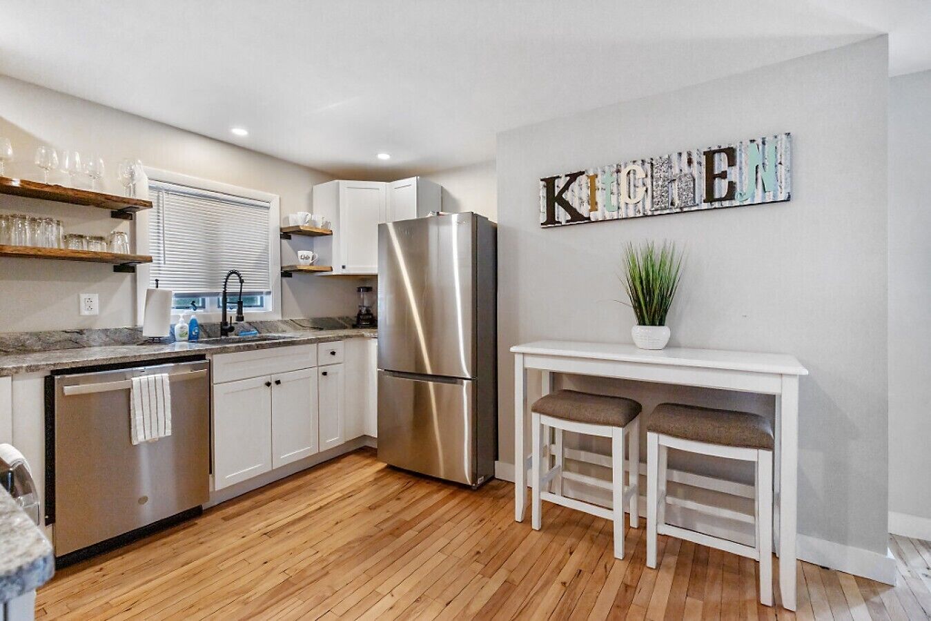 A modern kitchen with white cabinets, a stainless steel refrigerator, a wooden floor, and a bar area with stools.