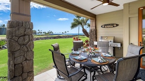 A covered outdoor dining area with a view of a well-maintained lawn and palm trees, a stone pillar, and a ceiling fan.